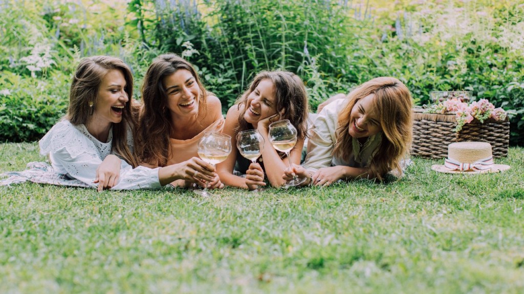 Women laying on the grass in a garden drinking wine and laughing, enjoying their #SustainableSummer.