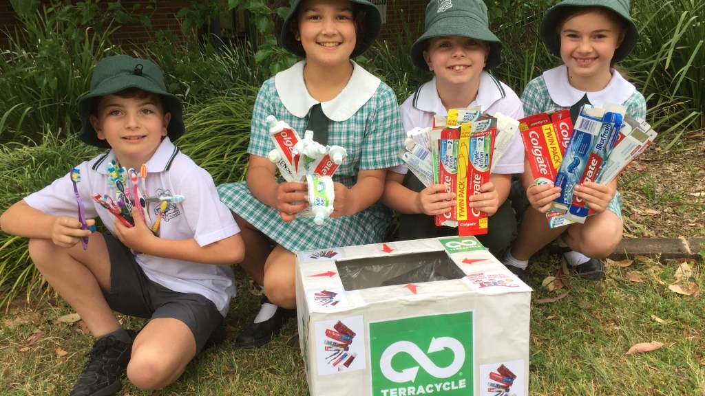 Kids in school uniforms holding toothbrushes and toothpaste tubes in front of a collection box, ready for recycling via TerraCycle