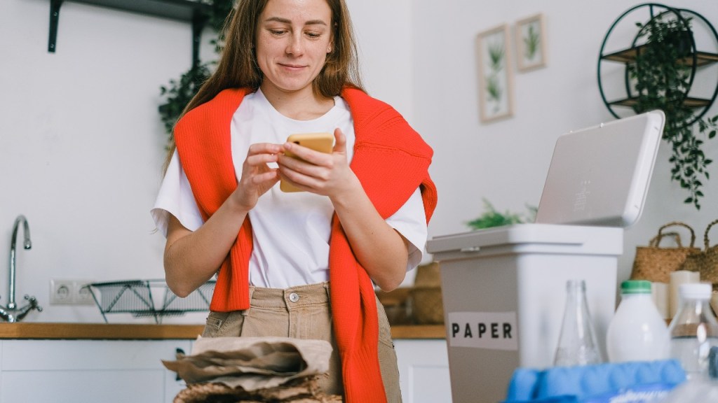 Woman with orange jumper on shoulders looking at phone surrounded by rubbish that she's sorting into recycling categories.